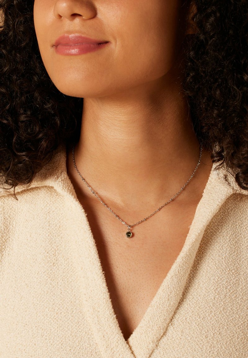 Zilveren ketting met een kleine groene hanger, gedragen op een beige textuur kledingstuk, rustend op het sleutelbeen.