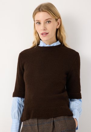 Blonde Frau trägt einen dunkelbraunen Kurzarm-Pullover über einem hellblau gestreiften Hemd und karierte Hosen, die Hände in den Taschen.