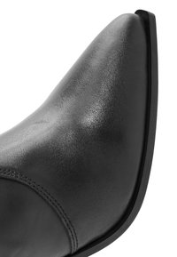 Zapato de cuero negro con un diseño de punta afilada, textura suave y un ligero tacón. Presenta costuras visibles a lo largo del costado y una suela de color contrastante.