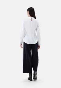 Weiße Bluse mit langen Ärmeln und einem Schlüsselloch-Rückendetail, kombiniert mit weiten dunkelblauen Hosen und schwarzen, knöchelhohen Stiefeln mit Absatz. Glatte Stofftextur.