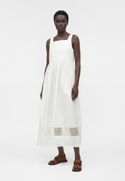 Γυναίκα με λευκό, μάξι φόρεμα χωρίς μανίκια με τετράγωνο ντεκολτέ και λεπτομέρεια με τρύπες πάνω από το τελείωμα, συνδυασμένο με καφέ σανδάλια με χαμηλό τακούνι, στέκεται μπροστά σε λευκό φόντο.