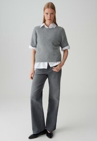 Vrouw in grijze korte mouwen trui over een wit shirt, grijze wijde jeans en zwarte puntige schoenen, staand tegen een eenvoudige achtergrond.