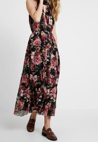 Robe maxi florale avec un fond noir, ornée de fleurs roses et blanches. Le tissu est léger et fluide, cintré à la taille par un lien. Des mocassins marron sont portés.