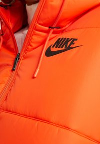 Ljus orange puffjacka med svart Nike-logga, dragkedja, textilier med struktur och elastisk dragsko. Har sydda sömmar för stabilitet.