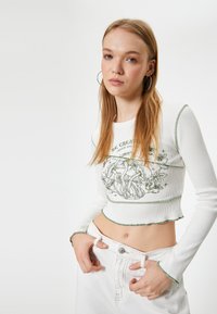 Witte geribbelde lange mouwen crop top met groene geborduurde details en geschulpte randen, met een botanische afbeelding op de voorkant.