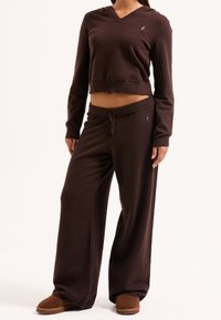 Felpa corta marrone con scollatura a V e tasca, abbinata a pantaloni marroni larghi con vita elasticizzata. Il modello indossa scarpe slip-on marroni.