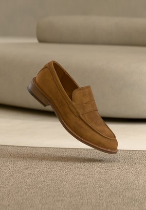 Brauner Wildleder-Herrenschuh im Mokassin-Stil schwebt über einem beigen Teppich vor einem passenden beigen Sofa im Hintergrund.