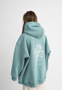 Felpa con cappuccio in misto cotone color turchese, caratterizzata da un disegno a linea bianca di un volto con accenti floreali sulla parte posteriore. Design morbido e comodo.