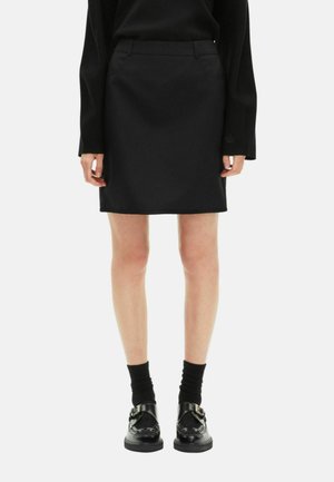 Black tailored mini skirt with a smooth texture, featuring side pockets and a waistband. Worn with black socks and shiny black shoes.