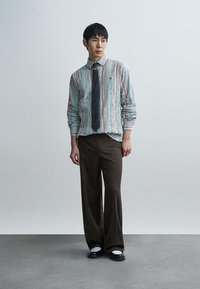 Μπλε και λευκό ριγωτό μακρυμάνικο πουκάμισο με κόκκινες λεπτομέρειες, συνδυασμένο με καφέ παντελόνι με φαρδιές μπότες και μαύρα παπούτσια με λευκές σόλες.