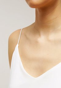 Camisole blanche avec de fines bretelles, un décolleté en V profond et une texture douce et lisse. Zone du cou et de la clavicule visible.
