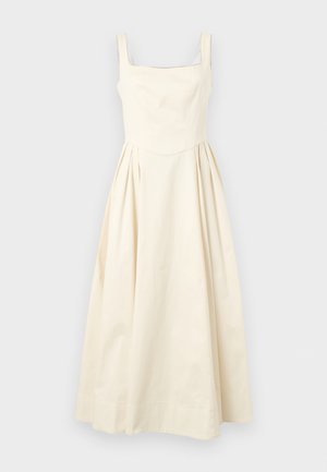 Nő krémszínű ujjatlan midi ruhában, sötét napszemüvegben és barna bojtos mokaszínben, egy kézzel a csípőjén áll.