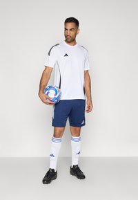 Λευκό αθλητικό μπλουζάκι με μαύρες ρίγες, ναυτικές σορτς, λευκές κάλτσες με μπλε ρίγες και μαύρα παπούτσια. Κρατάει μια πολύχρωμη μπάλα ποδοσφαίρου.