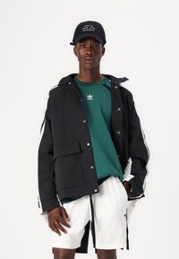 Μαύρο μπουφάν με λευκές ρίγες, πράσινο μπλουζάκι με λογότυπο και λευκές βερμούδες. Ένα καπέλο με κείμενο ολοκληρώνει την εμφάνιση, υπογραμμίζοντας την αθλητική ενδυμασία.