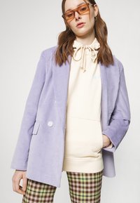 Blazer en velours côtelé lavande à deux boutons, porté sur un hoodie crème avec cordon de serrage. Associé à un pantalon à carreaux dans des tons de marron et de vert.