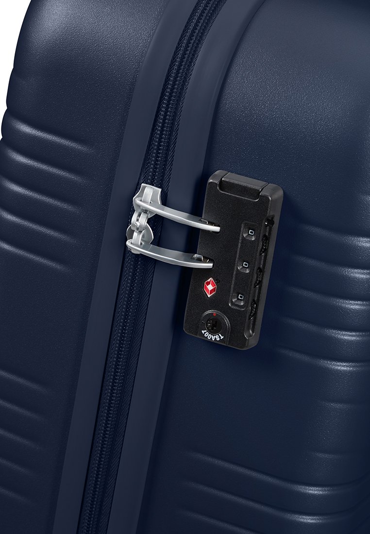 Valigia rigida blu navy con scanalature verticali, cerniera argentata e lucchetto a combinazione nero sul lato, dotato di tre dischi e un'etichetta di sicurezza.