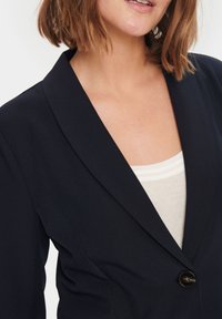 Jūras spēku bleizers ar iedobtu atlokšanu, vienu melnu pogu un strukturētu dizainu. Valkāts virs gaiši bežīgas ribotas blūzes. Gluda tekstūra.