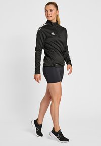 Zwarte zip-up sportjas met lange mouwen en logo; gecombineerd met zwarte shorts en zwarte sneakers met witte accenten.