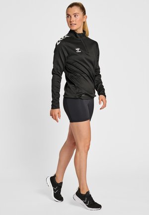 Zwarte zip-up sportjas met lange mouwen en logo; gecombineerd met zwarte shorts en zwarte sneakers met witte accenten.