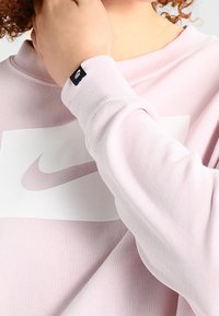 Světle růžová mikina s bílým logem Nike. Jemná tkanina, dlouhé rukávy, žebrované manžety a malá černá visačka na rukávu.