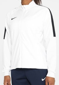 Weiße Zip-Jacke mit hohem Kragen, schwarzen Schulterstreifen und Nike-Logo. Glattes Material mit tailliertem Schnitt, kombiniert mit dunkelblauen Hosen.