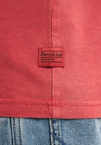 Camisa de algodón roja con una textura suave que cuenta con una etiqueta negra etiquetada "Petrol Ind" y jeans de mezclilla con costuras visibles.