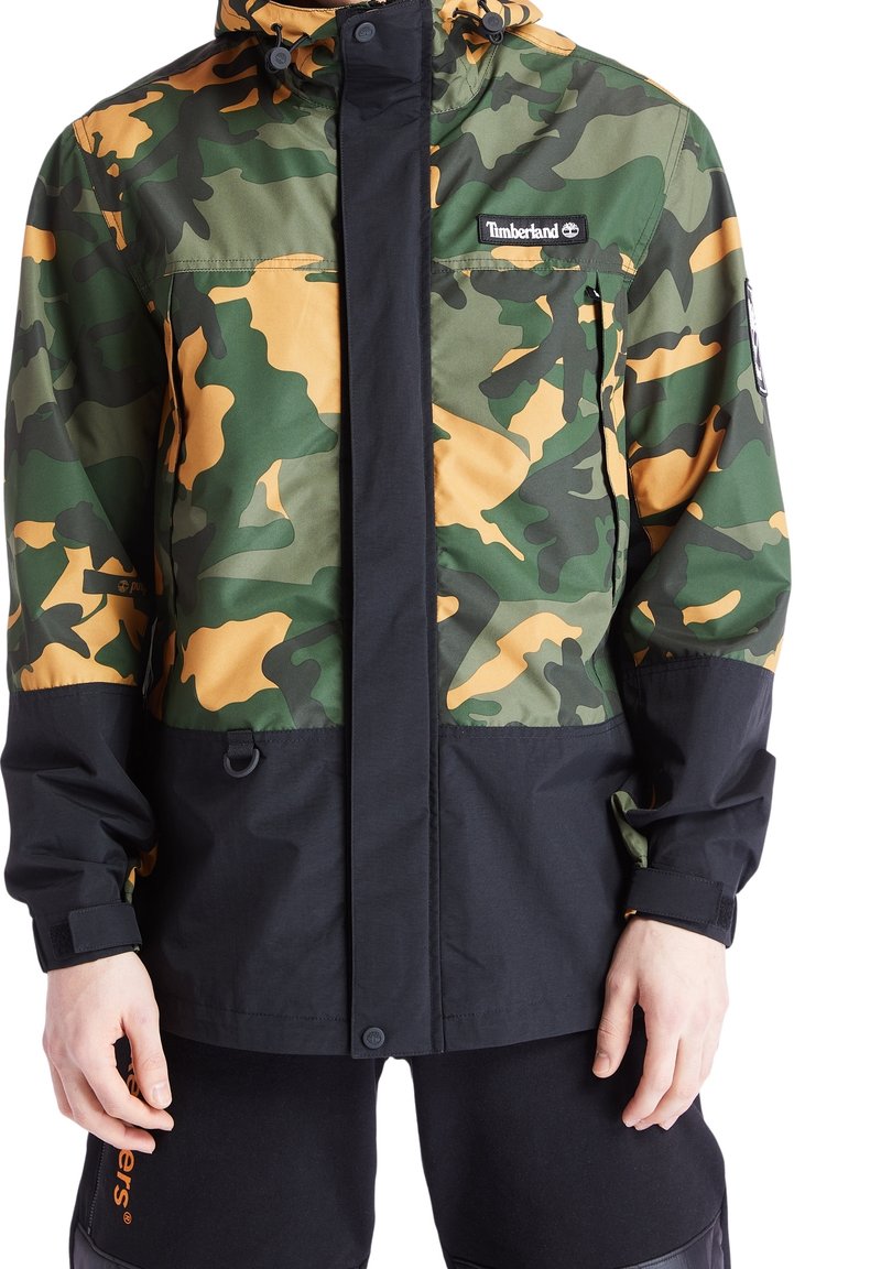 Kamouflagejacka i grön och orange, med en svart nederdel, utrustad med huva, dragkedjestängning och Timberland-logotyp på bröstet.