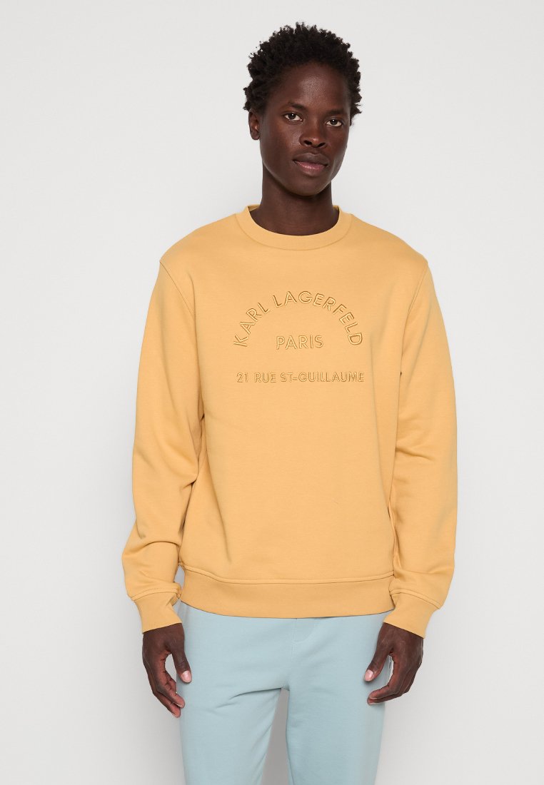 Junger Mann, der einen gelben Karl Lagerfeld Paris Sweatshirt und eine hellblaue Hose trägt, steht vor einem schlichten hellen Hintergrund.