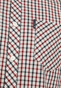 Camicia a quadri rossa, nera e bianca con chiusura a bottoni, dotata di taschino sul lato sinistro e un’etichetta cucita sulla tasca. Tessuto di cotone.