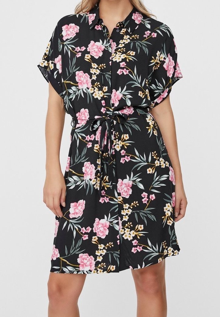 Femme portant une robe noire à manches courtes à fleurs roses, jaunes et blanches, avec des boutons sur le devant, un col et une ceinture à nouer à la taille.