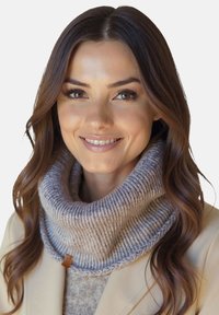 Smilende kvinde med langt, bølget brunt hår iført en beige frakke og gråt strikket højhalset tørklæde mod en enkel baggrund.