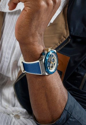 Chronograph watch with a blue textured silicone strap, silver casing, and blue dial featuring gold accents and multiple sub-dials.