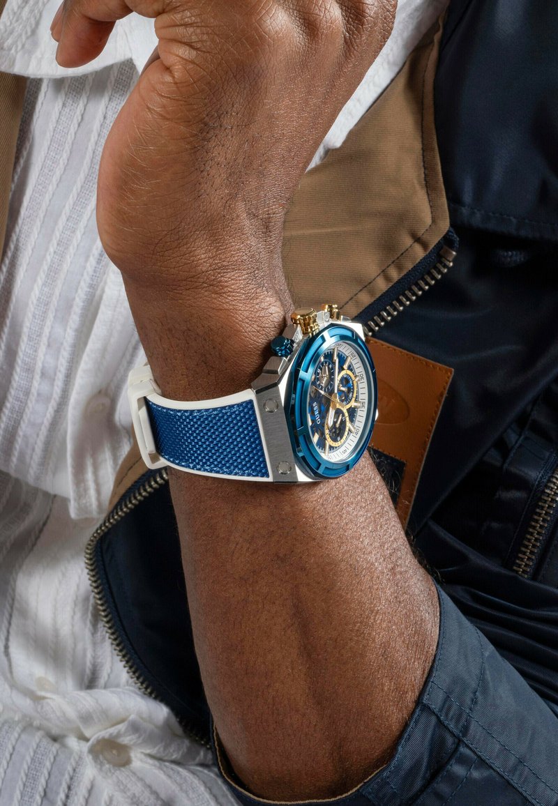 Blaues Chronographen-Armbanduhr mit einem Silber- und Goldgehäuse, blauen strukturierten Silikonarmband und einem weißen Zifferblatt mit goldenen Akzenten und Subzifferblättern.