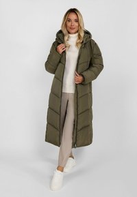 Nainen, joka on pukeutunut pitkään oliivinvihreään toppatakkiin valkoisen poolopaidan, vaaleiden housujen ja valkoisten tennareiden kanssa, seisoo yksinkertaista vaaleaa taustaa vasten.