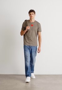 Tricou khaki cu un patch roșu, asociat cu blugi albaștri și adidași albi; culori simple și croială lejeră.