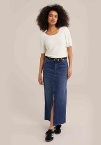 Džínová maxi sukně s rozparkem, spárovaná s texturovaným bílým topem. Černý opasek se stříbrnou sponou, na sobě má černé tenisky. Neutrální pozadí.