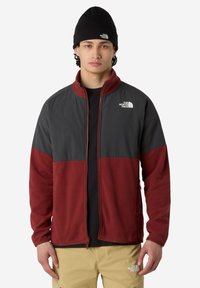Joven con un gorro negro, chaqueta con cremallera negra y burdeos, y pantalones beige, todos con el logo de The North Face.