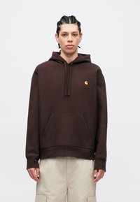 Felpa con cappuccio marrone in cotone, con tasca frontale, cappuccio con coulisse e un piccolo logo giallo. Indossata con pantaloni cargo beige.