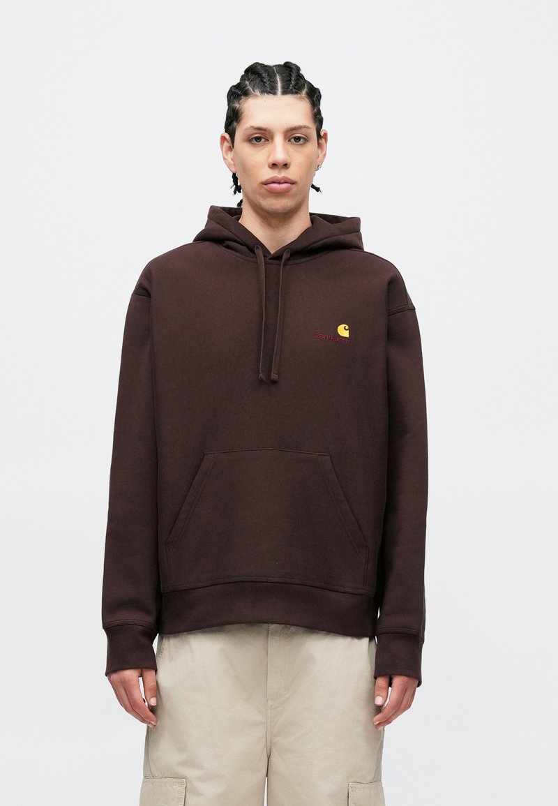 Brun hættetrøje lavet af bomuld, med forlomme, snøre i hætten og et lille gult logo. Båret sammen med beige cargobukser.