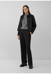 Vrouw staat gekleed in een zwarte cropped jack, grijze sweater met luipaardprint, zwarte wijde broek en zwarte plateau schoenen tegen een effen achtergrond.