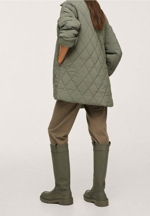 Personne portant une veste matelassée olive surdimensionnée, un pantalon marron et des bottes en caoutchouc vert olive montantes jusqu'aux genoux, debout les mains dans les poches.