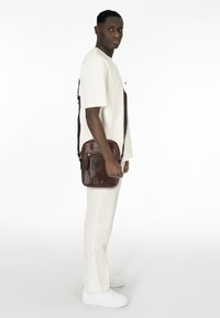 Brown leather messenger bag with a short strap, featuring a front zip pocket, rectangular shape, and smooth texture.