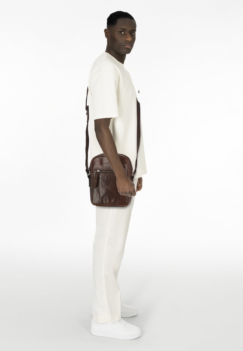Brown leather messenger bag with a short strap, featuring a front zip pocket, rectangular shape, and smooth texture.