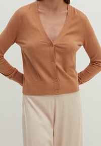 Jasnobrązowy kardigan, miękki dzianinowy materiał, dekolt w serek, siedem guzików z przodu, ściągacz na dole i przy mankietach, zestawiony z kremowymi spodniami.