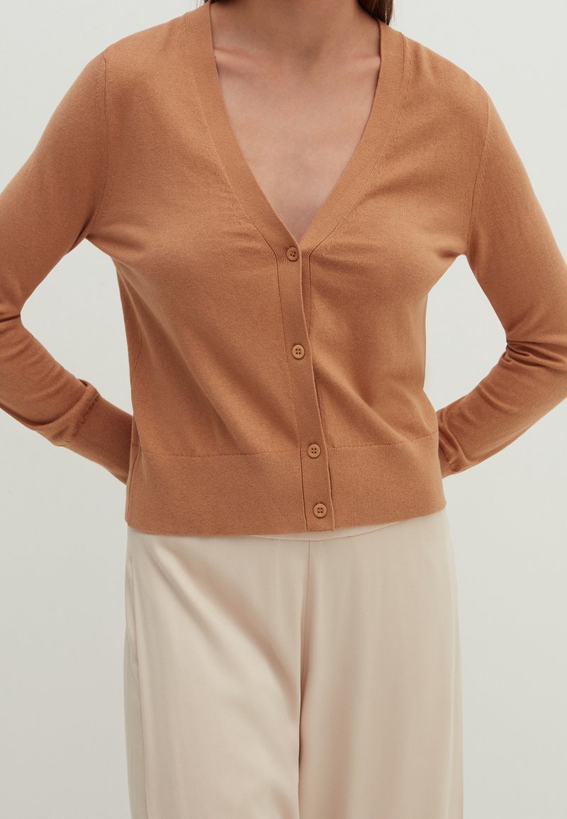 Jasnobrązowy kardigan, miękki dzianinowy materiał, dekolt w serek, siedem guzików z przodu, ściągacz na dole i przy mankietach, zestawiony z kremowymi spodniami.