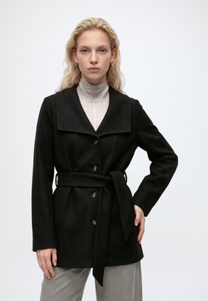 Jeune femme aux cheveux blonds porte un manteau noir ceinturé et un col roulé gris clair, debout devant un fond blanc uni.