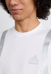 Bílý sportovní trik s stříbrnými popruhy, s jemným logem Adidas v odpovídající barvě. Hladká textura, přiléhavý střih, krátké rukávy.