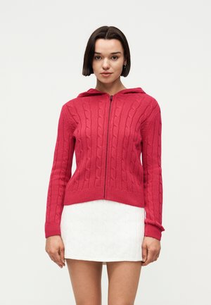 Νέα γυναίκα με κοντά σκούρα μαλλιά, που φοράει κόκκινο πλεκτό hoodie με φερμουάρ και λευκή υφασμάτινη μίνι φούστα, στέκεται μπροστά σε απλό φόντο.