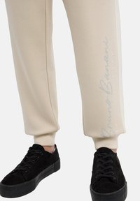Beiga sweatpants med slim fit, som har en vertikal silverdesign på sidan. Kombination med svarta mockaskor och texturerade svarta sulor.