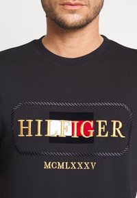 Svart bomulls-t-shirt med en kraftfull broderad "HILFIGER"-logotyp med gulddetaljer, omgiven av en repdesign; inkluderar "MCMLXXXV" under.